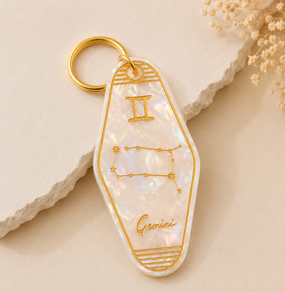 Keychain with Gemini zodiac design on a beige stone surface with dried flowers.