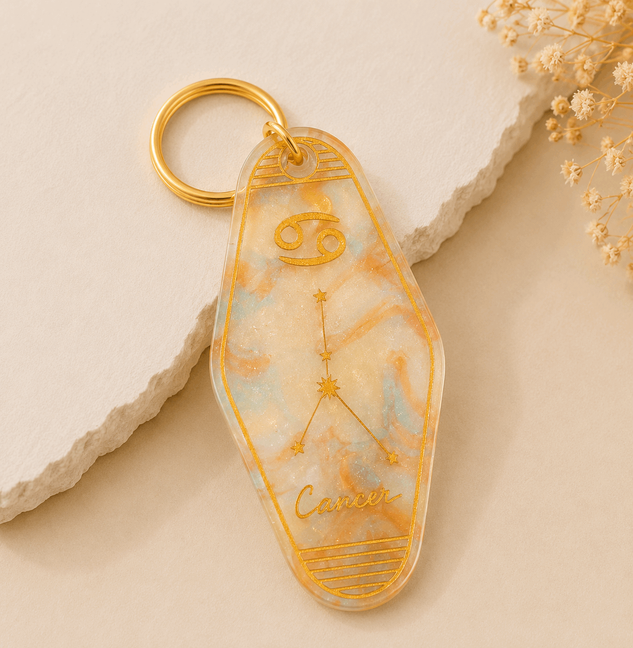 Keychain with Cancer zodiac design on a beige stone surface with dried flowers.