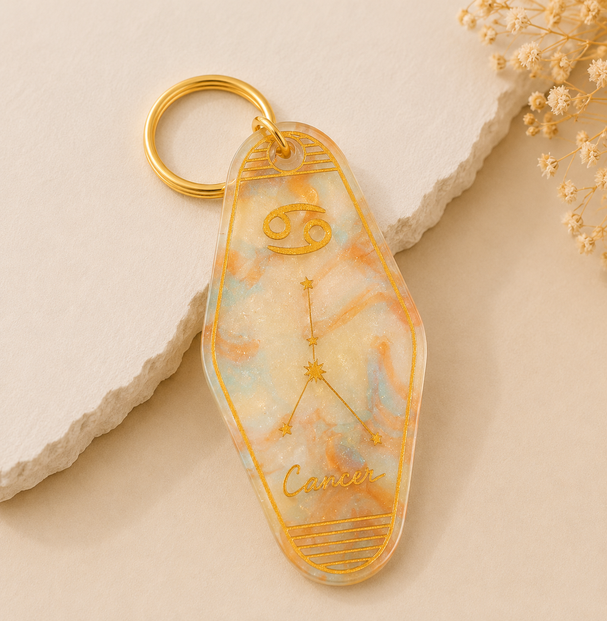 Keychain with Cancer zodiac design on a beige stone surface with dried flowers.