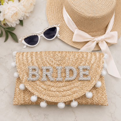 Straw clutch with 'BRIDE' embellishments, sun hat, and sunglasses on a light background
