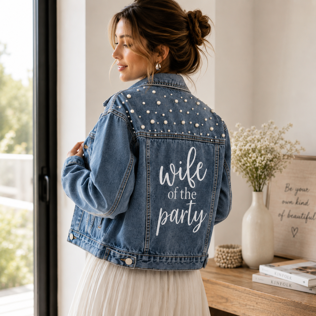 Woman wearing a denim jacket with 'wife of the party' text, standing in a bright room.