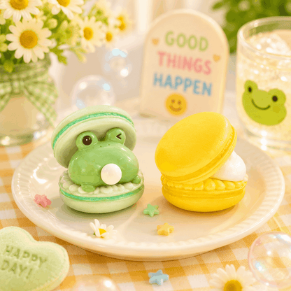 Two frog-shaped macarons on a plate with decorative elements and a 'Good Things Happen' sign in the background.
