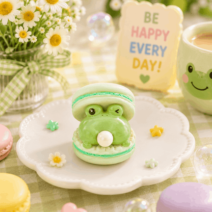 Green frog-shaped macaron on a white plate with flowers and a 'Be Happy Every Day' sign in the background.