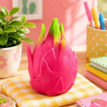 Pink dragon fruit-shaped object on a checkered cloth with a plant and stationery items in the background.