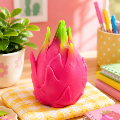 Pink dragon fruit-shaped object on a checkered cloth with a plant and stationery items in the background.