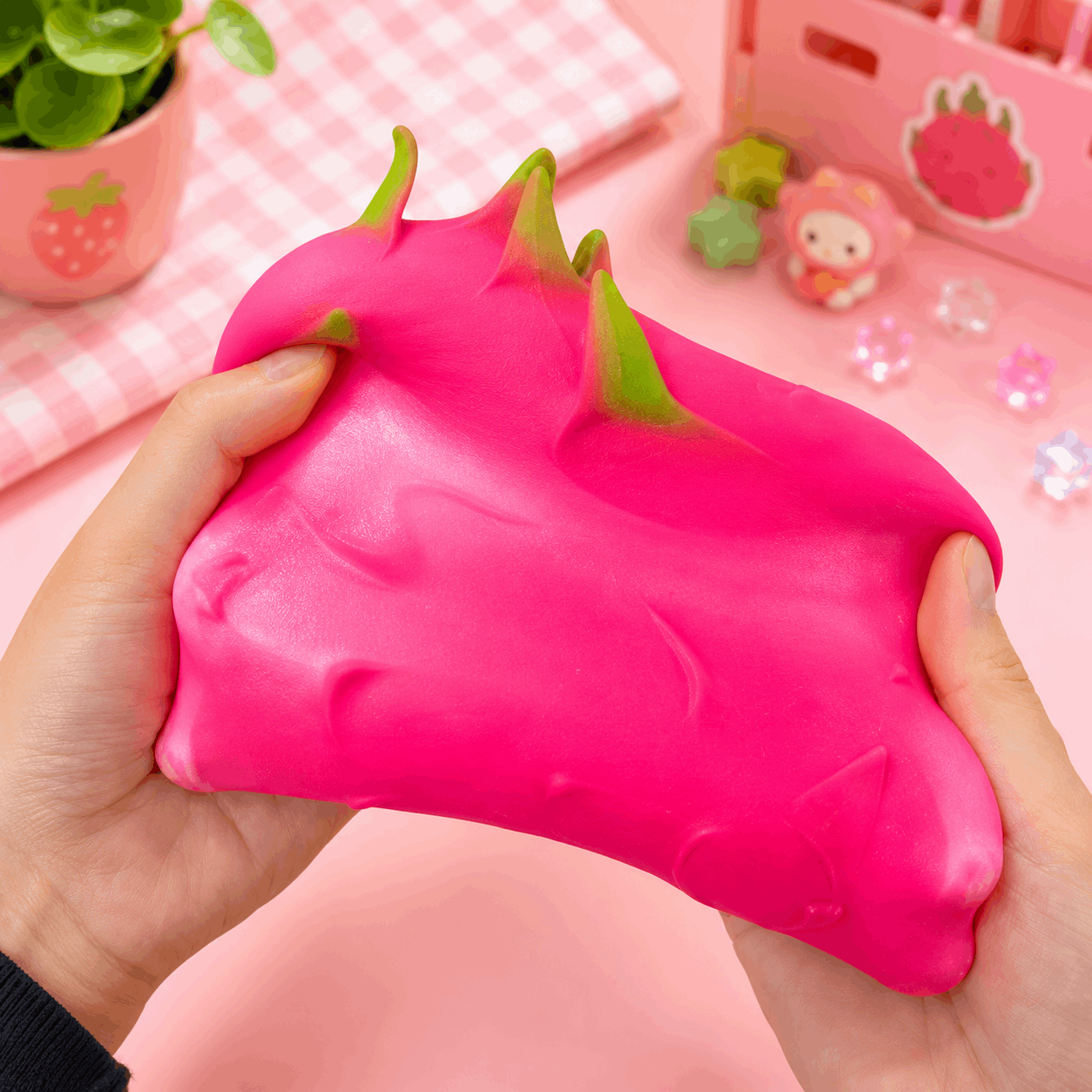 Pink dragon fruit-shaped squishy toy held in hands with a pink background