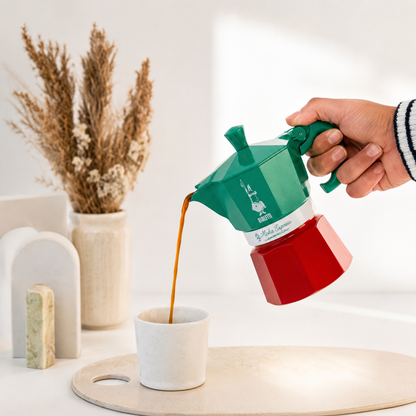 Green and red espresso maker being used to pour coffee into a white cup on a light wooden surface.