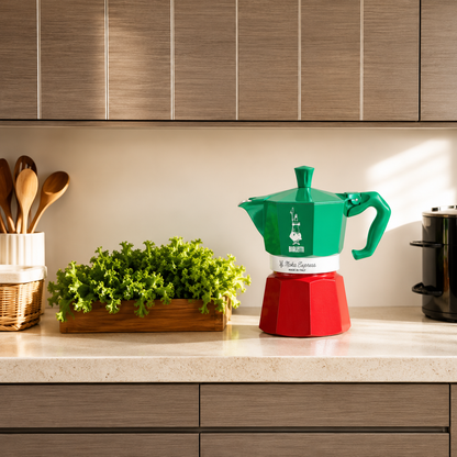 Green and red coffee maker on a kitchen counter with plants and utensils.