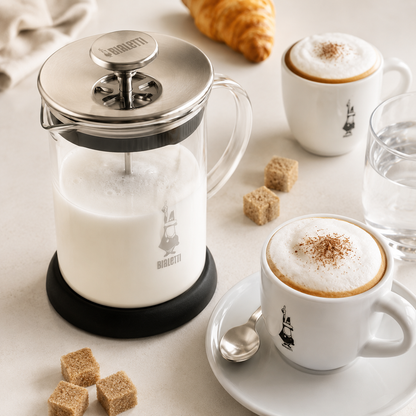 French press filled with milk, two cups of cappuccino, and a croissant on a light surface.