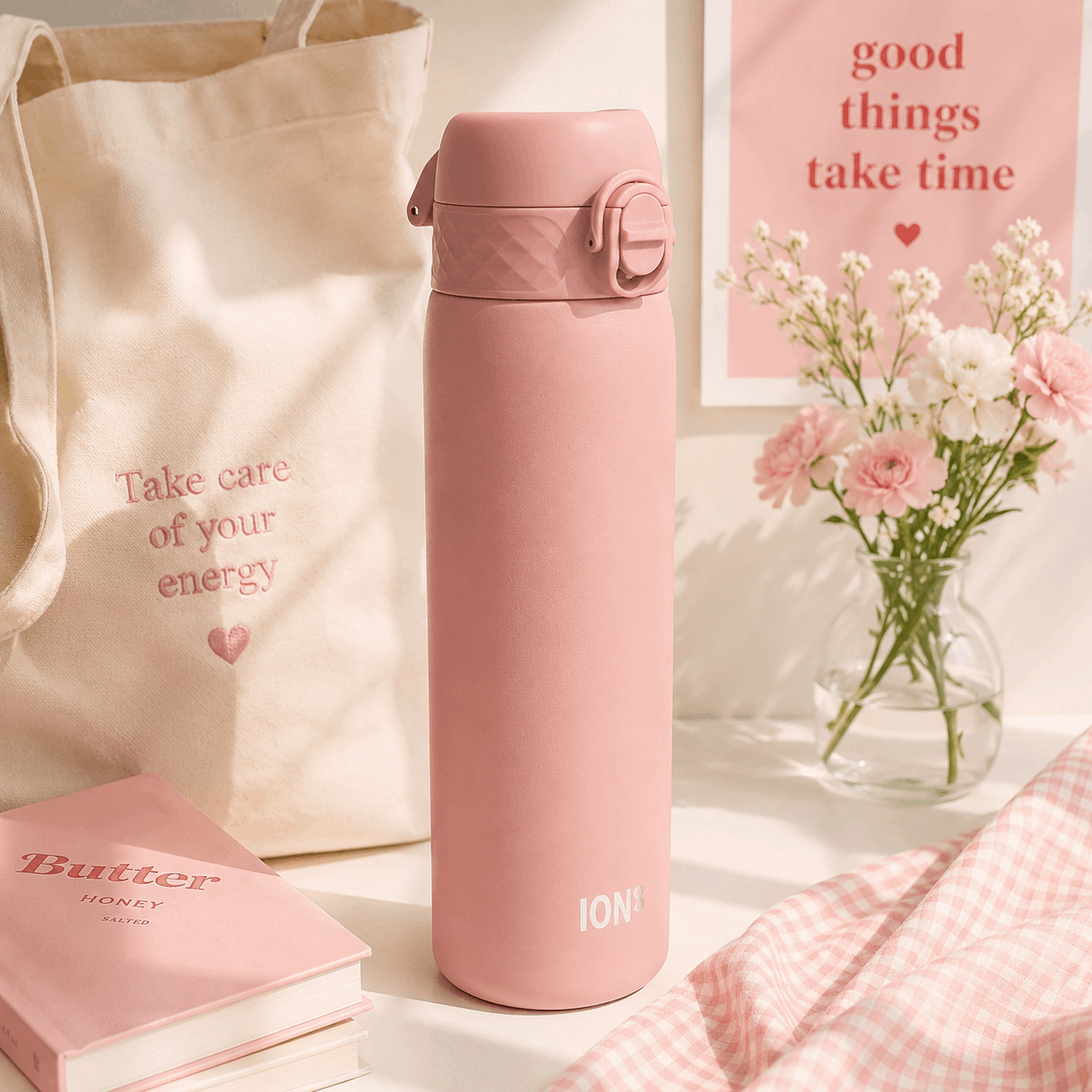 A pink insulated water bottle with a flip-top lid stands on a tabletop beside a cream canvas tote bag, a pink book, and a small vase of flowers. The bottle has the brand name ION+ printed near the bottom.