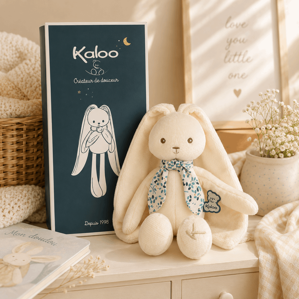 A cream-colored plush bunny toy with a blue floral bow sits next to its dark blue packaging box on a light wooden surface, styled in a warm, cozy nursery setting with a basket, blanket, and small potted flowers.