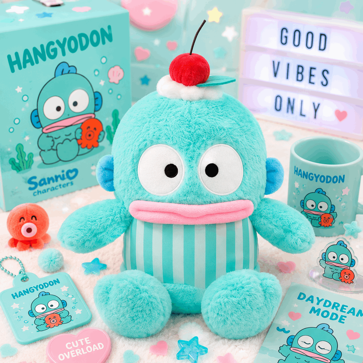 A Sanrio Hangyodon plush toy in bright aqua blue with a pink mouth and a red cherry on top, displayed with branded merchandise including a box, mug, keychain, acrylic charm, and printed cards on a pastel studio set.