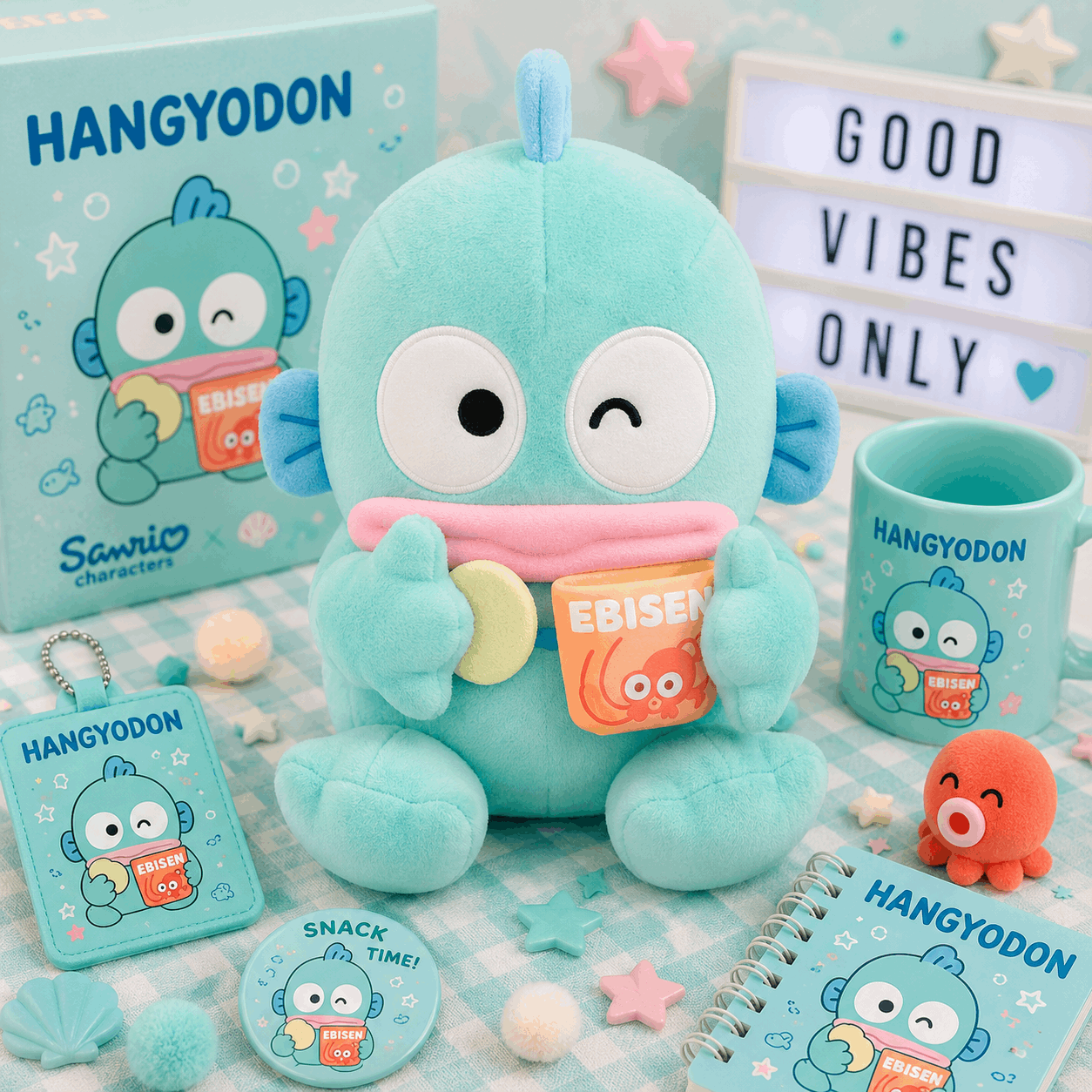 A Sanrio Hangyodon plush toy displayed with matching merchandise including a mug, notebook, card, and small accessory on a pastel checkered surface. The scene includes decorative props and a light box reading 'GOOD VIBES ONLY'.