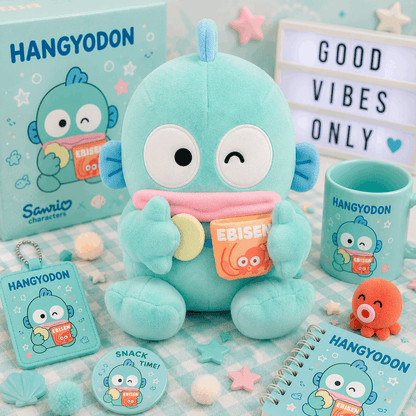 A Sanrio Hangyodon plush toy displayed with matching merchandise including a mug, notebook, card, and small accessory on a pastel checkered surface. The scene includes decorative props and a light box reading 'GOOD VIBES ONLY'.