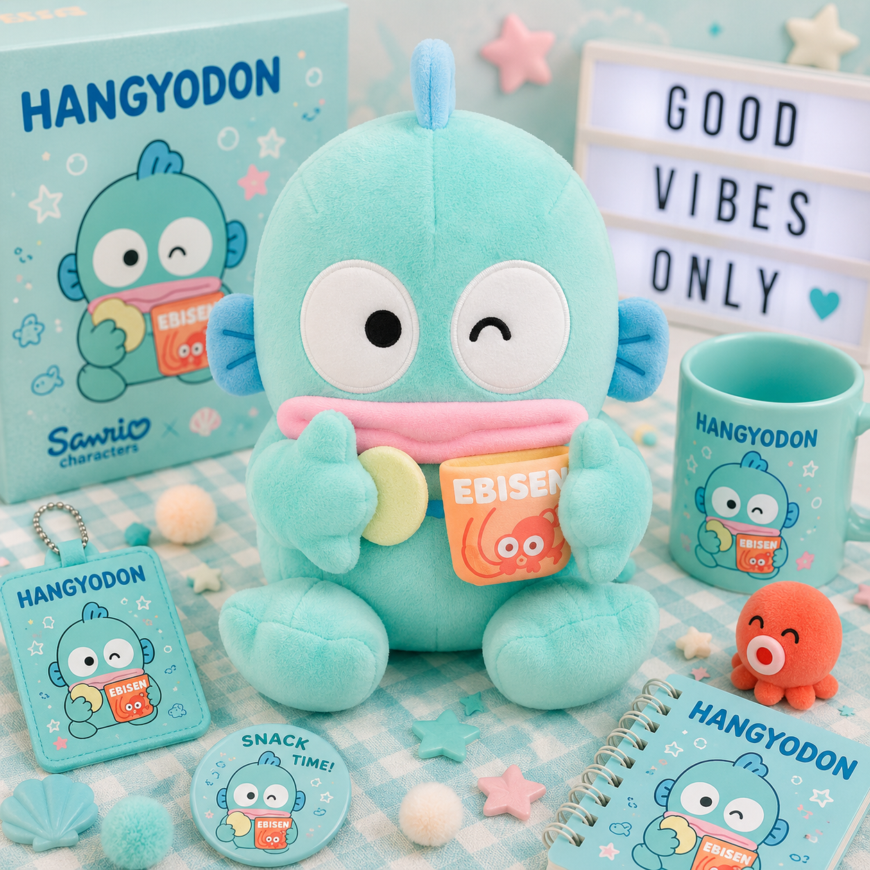 A Sanrio Hangyodon plush toy displayed with matching merchandise including a mug, notebook, card, and small accessory on a pastel checkered surface. The scene includes decorative props and a light box reading 'GOOD VIBES ONLY'.