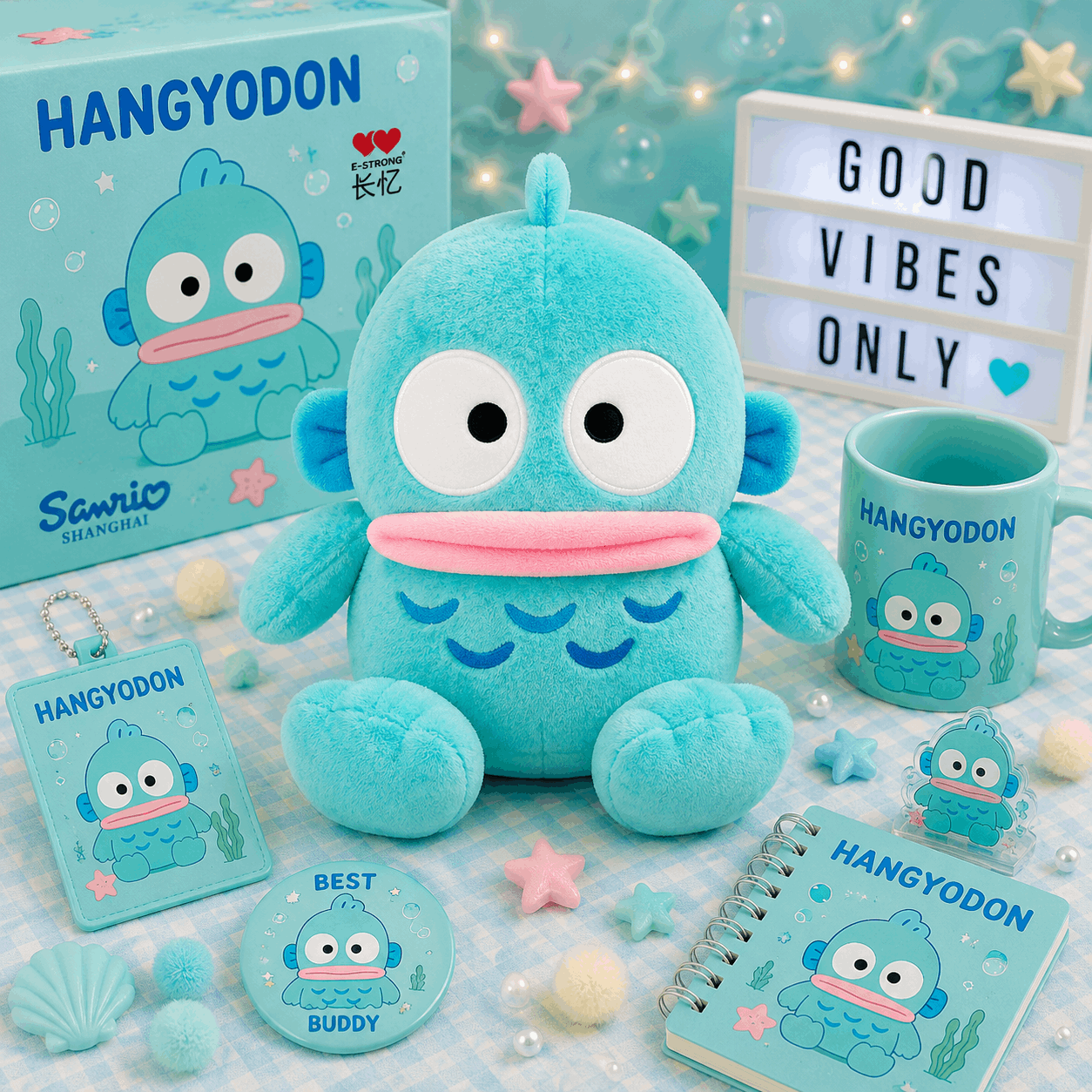 A colorful product display featuring a light blue Hangyodon plush toy, a matching mug, a notebook, a keychain, a round pin, and a character box on a checkered blue and white blanket with star and bubble decorations.