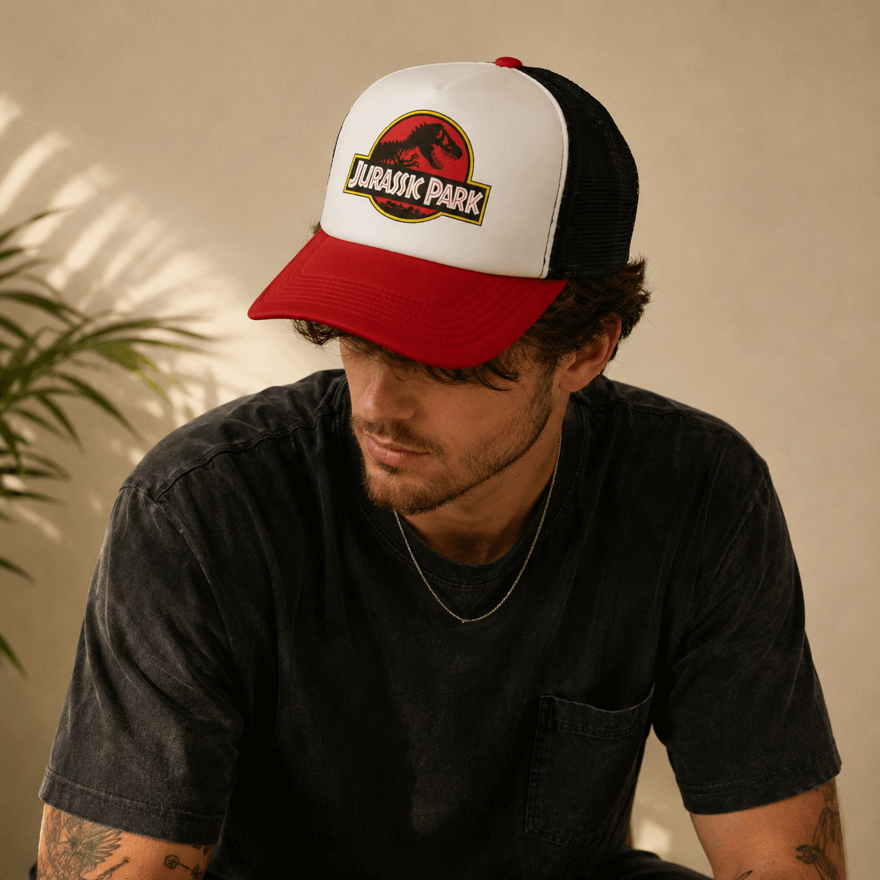 A man wearing a red and white Jurassic Park trucker hat with a black mesh back, looking down in a warm, sunlit setting.