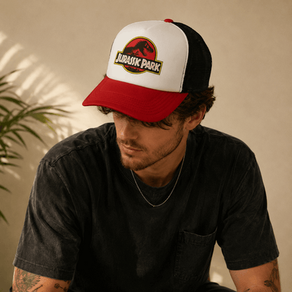 A man wearing a red and white Jurassic Park trucker hat with a black mesh back, looking down in a warm, sunlit setting.
