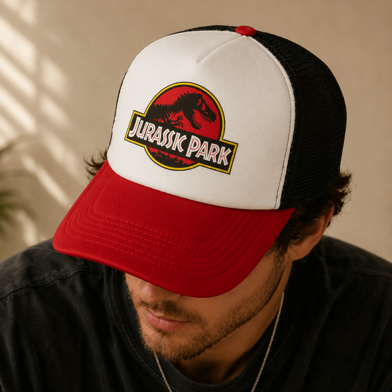 A man wearing a red, white, and black Jurassic Park trucker hat, looking down. The hat features the iconic red and black Jurassic Park T-Rex logo on the front white panel.
