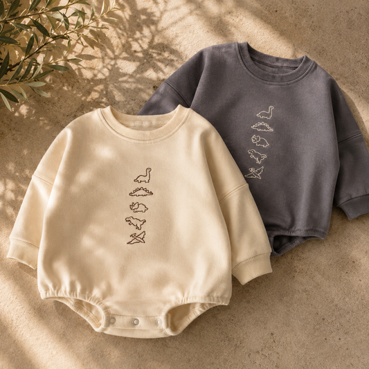 Two baby long-sleeve bodysuits laid flat on a textured beige surface with soft leaf shadows. The front bodysuit is cream with a vertical line of small outlined bird motifs, and the back bodysuit is dark gray with a similar motif.