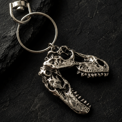 A silver metal keychain featuring two dinosaur skull charms, one larger and one smaller, attached to a split key ring, displayed on a dark textured slate background.