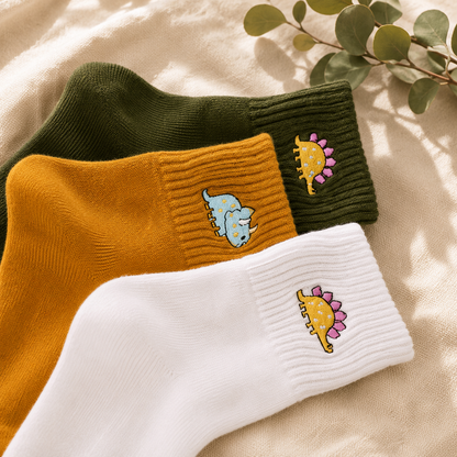 Three ribbed ankle socks arranged in an overlapping flat lay: one dark olive green, one mustard yellow, and one white. Each sock has a small embroidered dinosaur patch on the cuff.