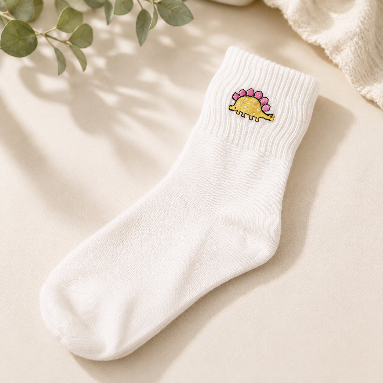 A single white ribbed ankle sock with a small embroidered yellow dinosaur with pink spikes on the cuff, displayed on a warm beige surface with soft shadows and decorative green leaves.