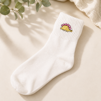 A single white ribbed ankle sock with a small embroidered yellow dinosaur with pink spikes on the cuff, displayed on a warm beige surface with soft shadows and decorative green leaves.
