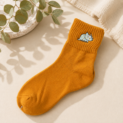 A single mustard yellow ribbed ankle sock with a small embroidered light blue dinosaur on the cuff, styled on a beige surface with soft shadows, a plant leafy sprig, and a cream knit fabric.