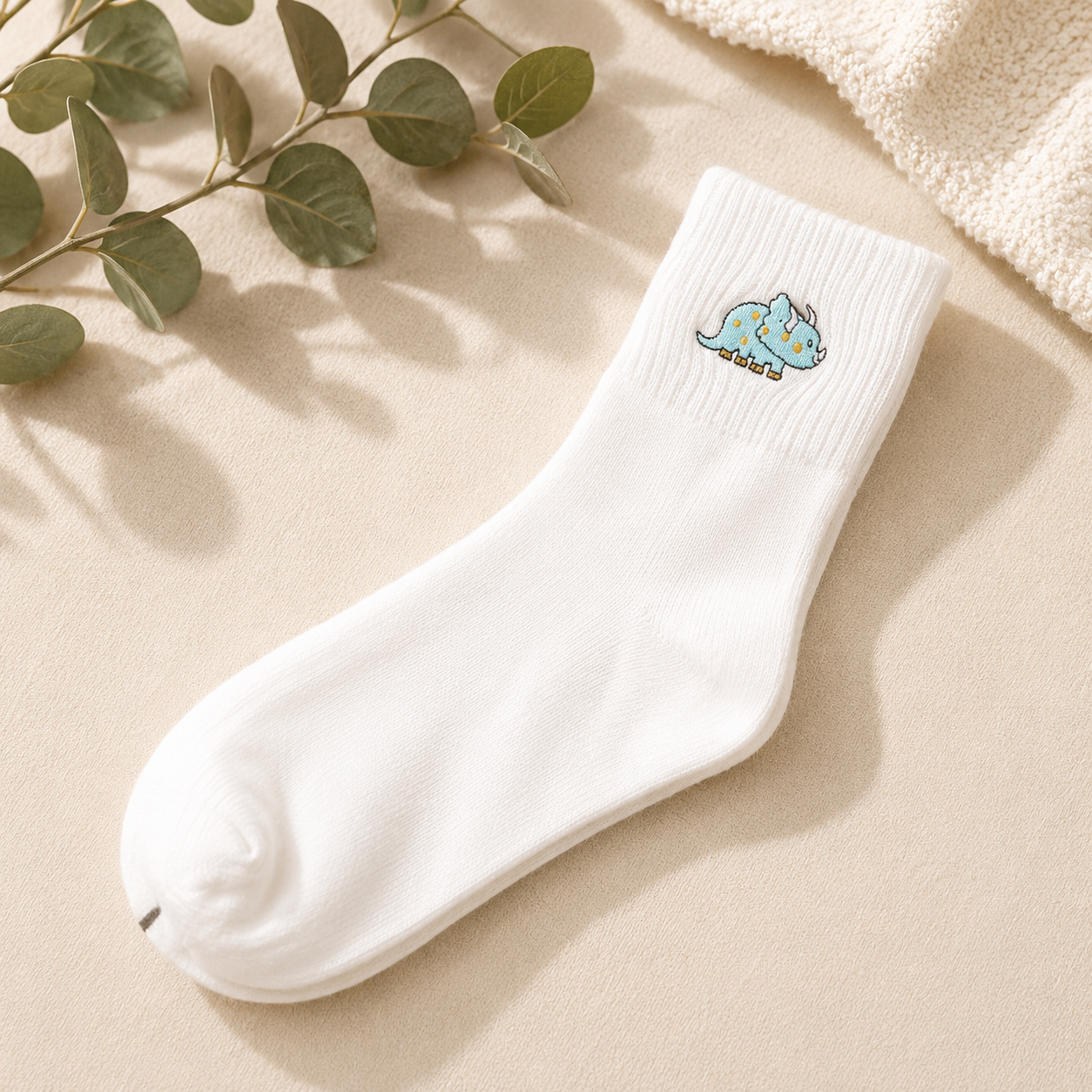 A single white crew sock with a small embroidered blue dinosaur on the cuff, styled on a beige fabric surface with green leaves and a cream knit textile in the background.