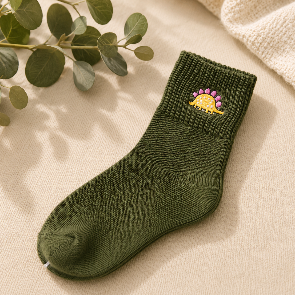 An olive green ribbed ankle sock with a small embroidered dinosaur design on the cuff, styled on a beige textured fabric with eucalyptus leaves.