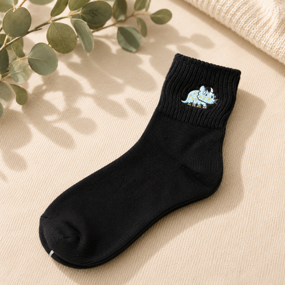 A single black crew sock with a small embroidered cartoon dinosaur on the cuff, styled on a beige textured fabric with eucalyptus leaves and a cream knit blanket.