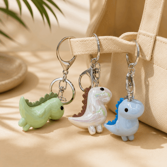 Three small dinosaur-shaped keychains with silver key rings are displayed on a beige fabric surface next to a canvas tote bag. The keychains are pastel green, white with iridescent pink detailing, and light blue with a blue back.
