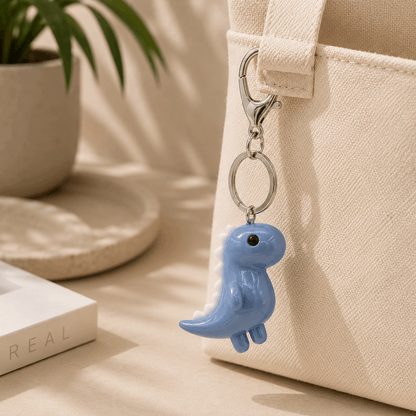 A small glossy blue dinosaur keychain charm with a silver clasp, hanging from a cream canvas tote bag in a softly lit indoor setting with a plant and book in the background.