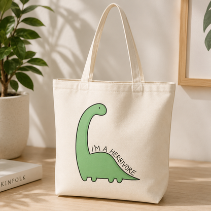An off-white canvas tote bag with a green cartoon dinosaur and the text 'I'M A HERIVORE' printed on the front, displayed on a wooden table next to a potted plant.