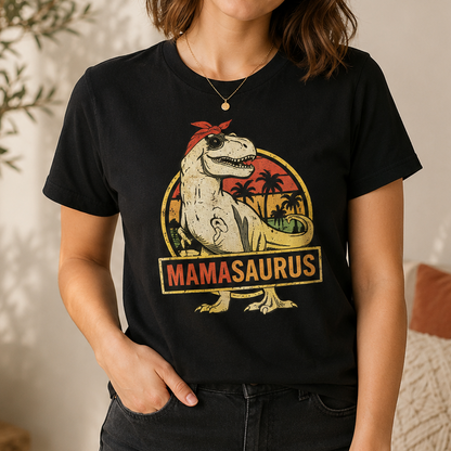 A woman wearing a black graphic t-shirt featuring a retro-style dinosaur design with the text 'MAMASAurus'. The t-shirt has a distressed yellow and red graphic of a dinosaur in a tropical scene, and the model is wearing dark jeans in a softly lit indoor setting.