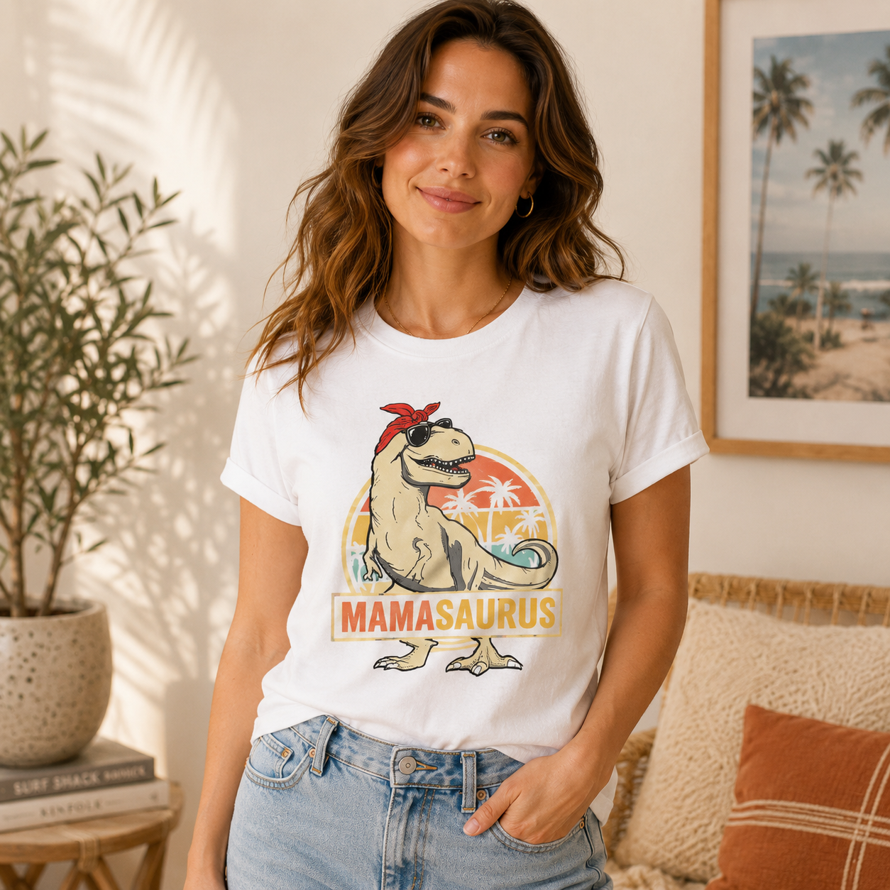 A woman wearing a white graphic t-shirt with a retro-style 'MAMASAUROUS' dinosaur design, paired with light-wash blue jeans, standing in a cozy, sunlit room with a potted plant and framed beach art in the background.