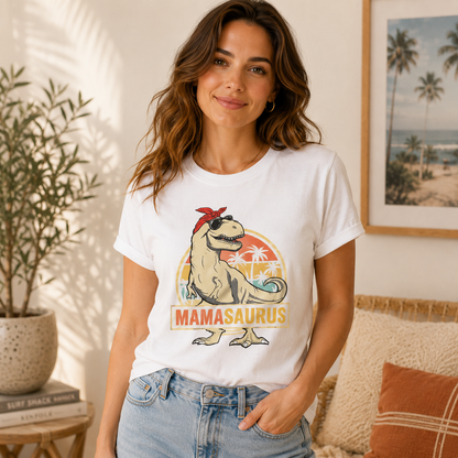 A woman wearing a white graphic t-shirt with a retro-style 'MAMASAUROUS' dinosaur design, paired with light-wash blue jeans, standing in a cozy, sunlit room with a potted plant and framed beach art in the background.