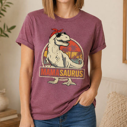 A woman wearing a heathered mauve-purple t-shirt with a retro-style graphic of a white T-Rex wearing sunglasses and a red bandana, with the text 'MAMA SAURUS' below. She is also wearing blue jeans in a cozy indoor setting.