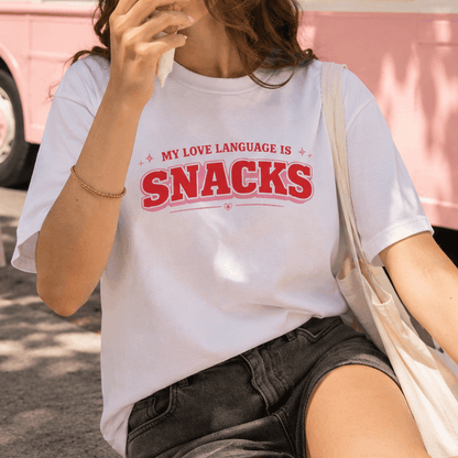 A woman wearing a white graphic T-shirt with the text 'MY LOVE LANGUAGE IS SNACKS' in red and pink, paired with dark denim shorts and a cream tote bag, set against a pink vehicle background.