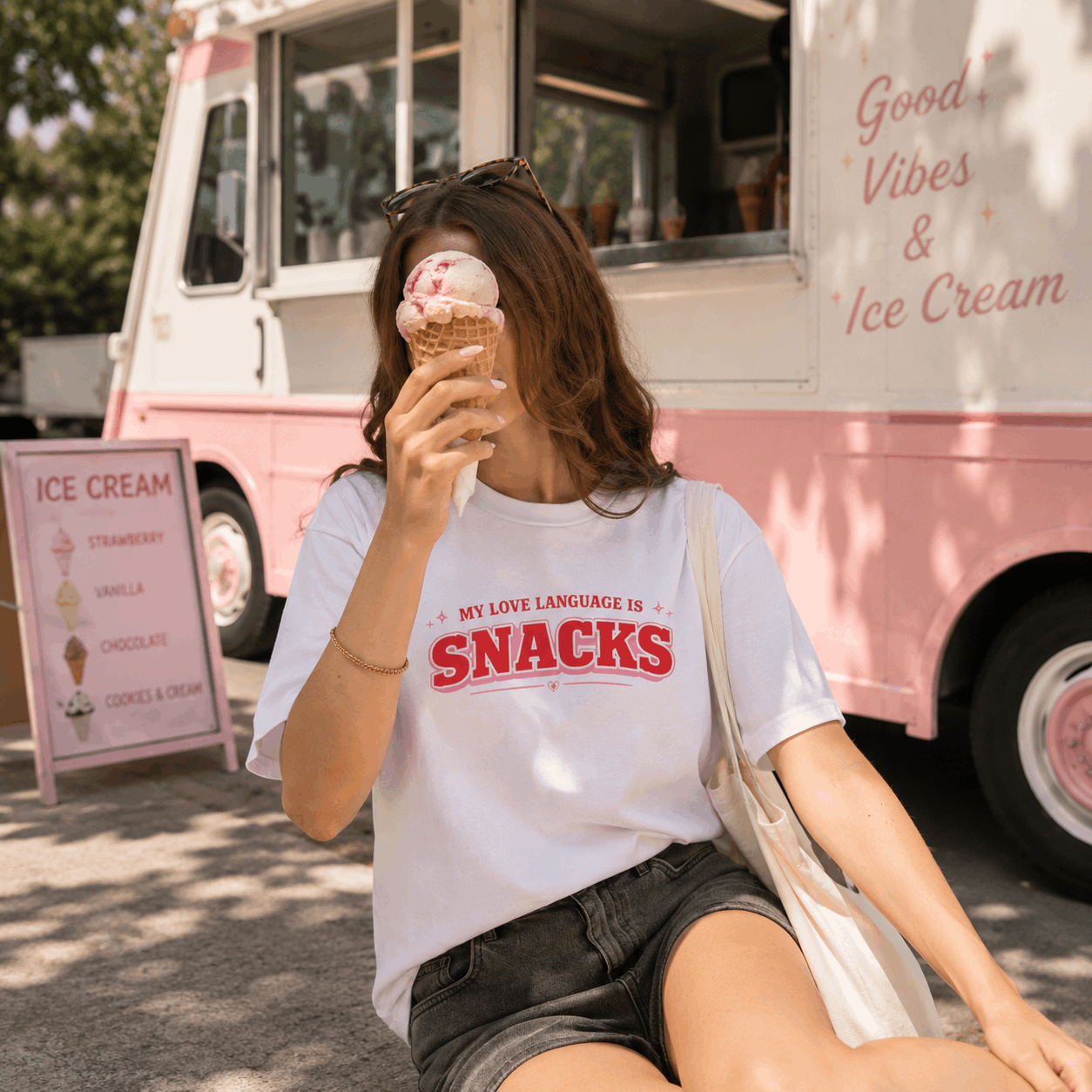A woman sitting outdoors in front of a pink ice cream truck, wearing a white graphic T-shirt with red text that reads 'MY LOVE LANGUAGE IS SNACKS' and holding an ice cream cone.