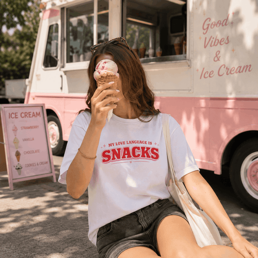 A woman sitting outdoors in front of a pink ice cream truck, wearing a white graphic T-shirt with red text that reads 'MY LOVE LANGUAGE IS SNACKS' and holding an ice cream cone.