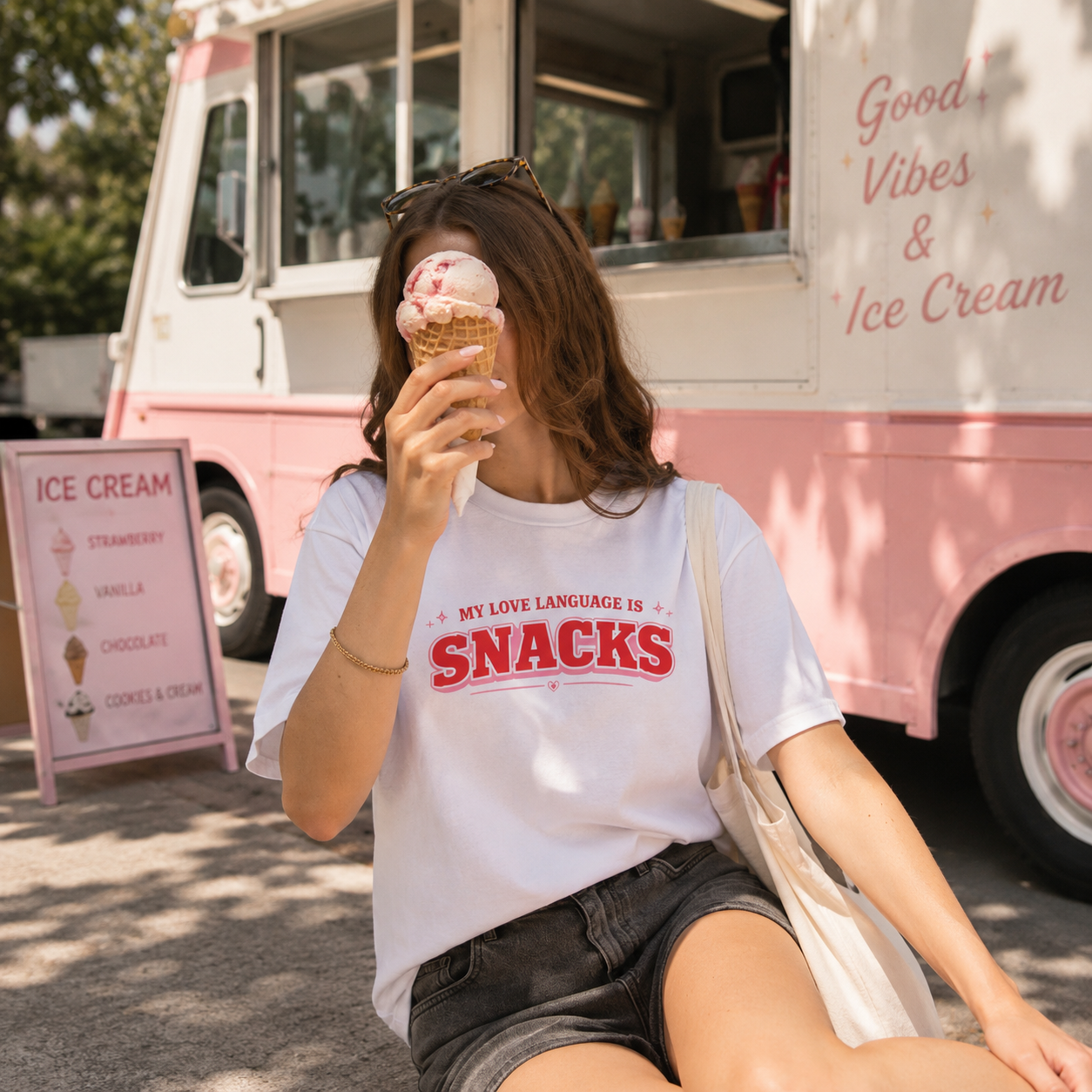 A woman sitting outdoors in front of a pink ice cream truck, wearing a white graphic T-shirt with red text that reads 'MY LOVE LANGUAGE IS SNACKS' and holding an ice cream cone.