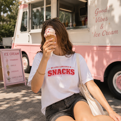 A woman sitting outdoors in front of a pink ice cream truck, wearing a white graphic T-shirt with red text that reads 'MY LOVE LANGUAGE IS SNACKS' and holding an ice cream cone.