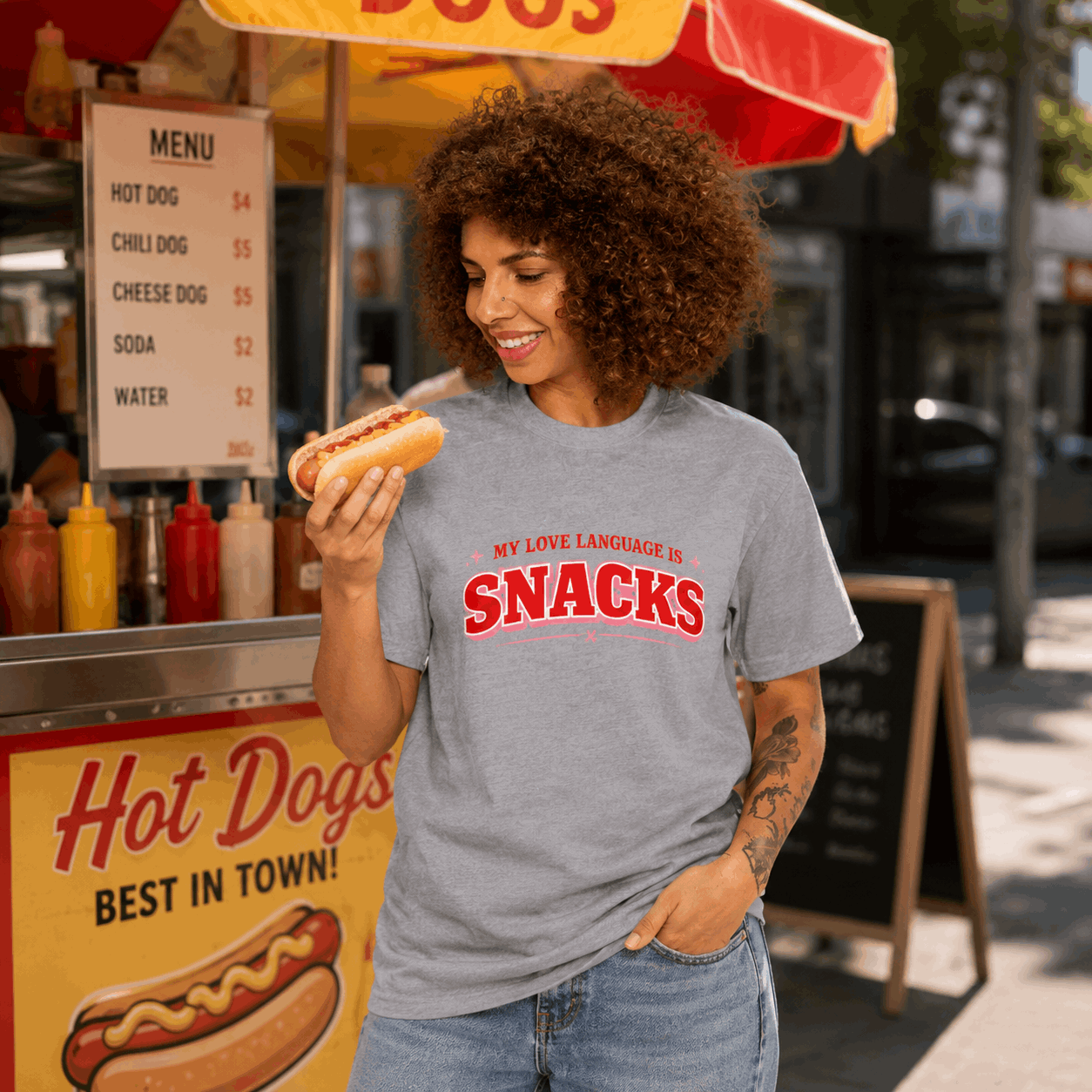 A woman with curly hair stands at a hot dog stand wearing a heather gray t-shirt with 'MY LOVE LANGUAGE IS SNACKS' printed in red across the chest. She is holding a hot dog and smiling, with the food cart and street background visible.