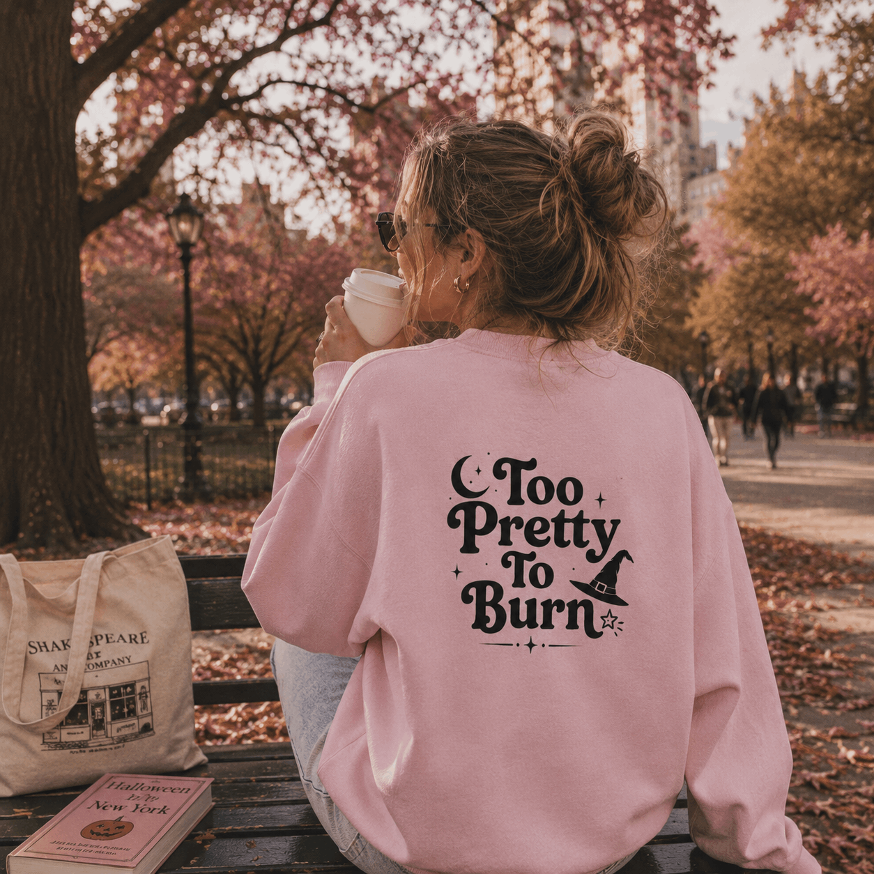 A woman sitting on a park bench with her back to the camera, wearing a light pink sweatshirt with the black text 'Too Pretty To Burn' and a witch hat graphic on the back. She is holding a white coffee cup, with an autumn park scene in the background.