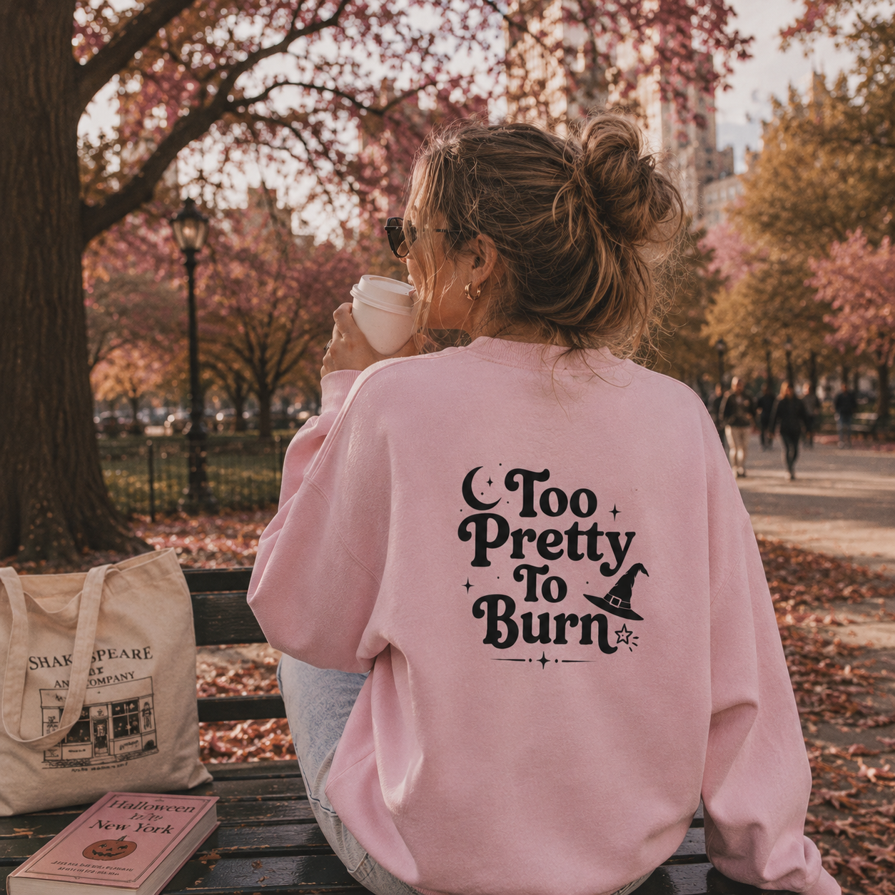 A woman sitting on a park bench with her back to the camera, wearing a light pink sweatshirt with the black text 'Too Pretty To Burn' and a witch hat graphic on the back. She is holding a white coffee cup, with an autumn park scene in the background.