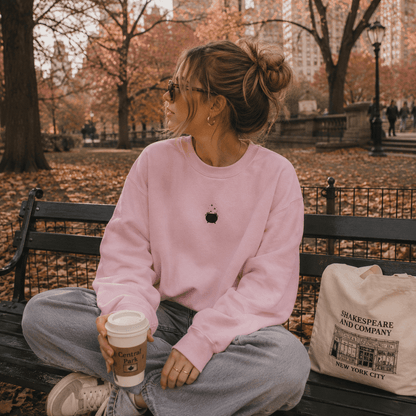 A woman sits on a park bench wearing a light pink crewneck sweatshirt with a small black apple logo, gray sweatpants, and holding a coffee cup. A canvas tote bag with 'Shakespeare and Company New York City' text rests beside her.