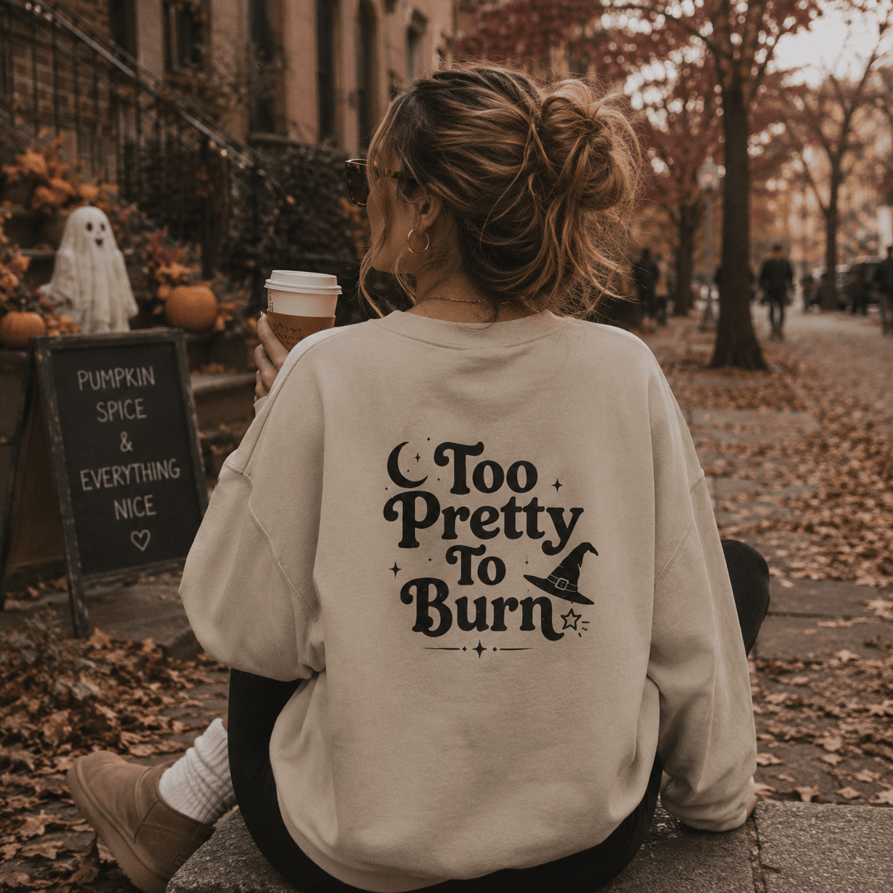 A woman sitting outdoors on a stone path in autumn, viewed from behind, wearing a cream-colored sweatshirt with a black graphic that reads 'Too Pretty To Burn' with a witch hat illustration. She holds a coffee cup, with pumpkins, a ghost decoration, and a cafe chalkboard sign visible in the background.