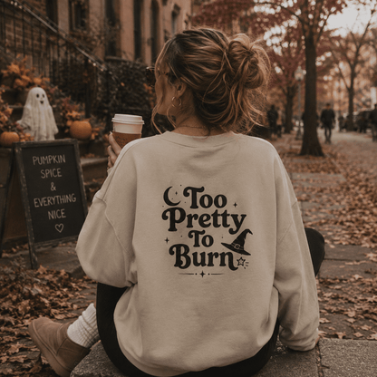A woman sitting outdoors on a stone path in autumn, viewed from behind, wearing a cream-colored sweatshirt with a black graphic that reads 'Too Pretty To Burn' with a witch hat illustration. She holds a coffee cup, with pumpkins, a ghost decoration, and a cafe chalkboard sign visible in the background.